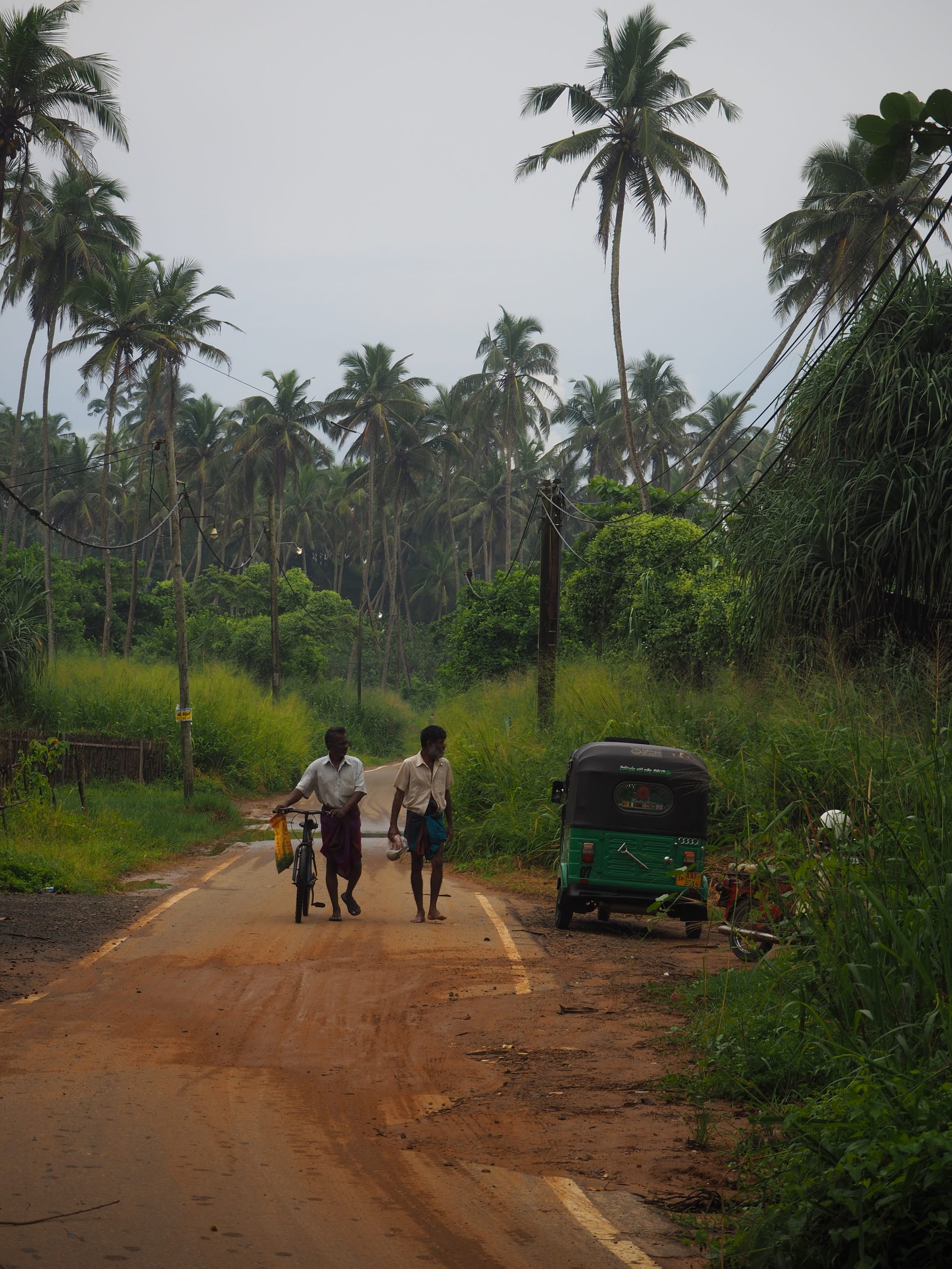 Two Sri Lankan men walking with a bike, beside a passing tuktuk