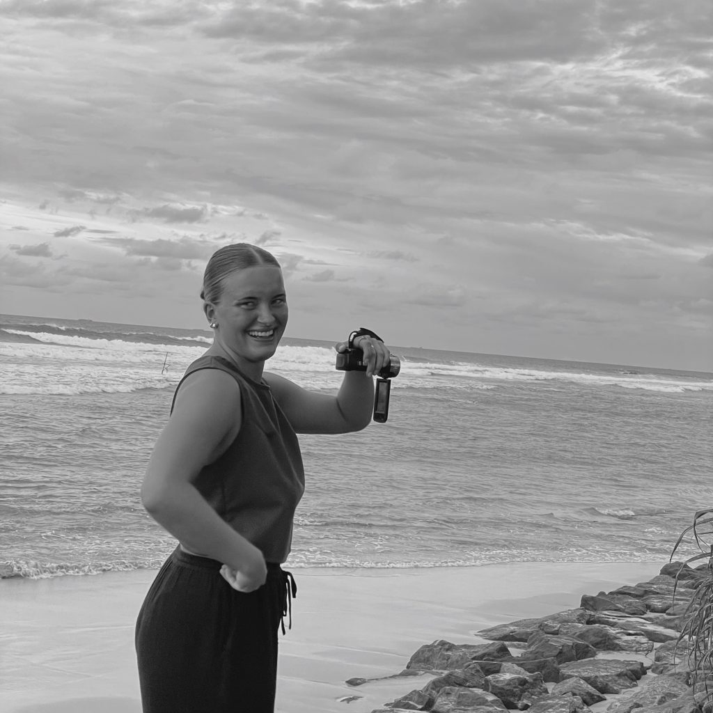 Girl smiling at the camera, holding a camcorder while recording the ocean