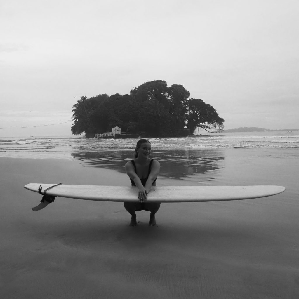 Girl balancing on her tippy toes on the beach, with a surfboard on her lap and an island in the distance