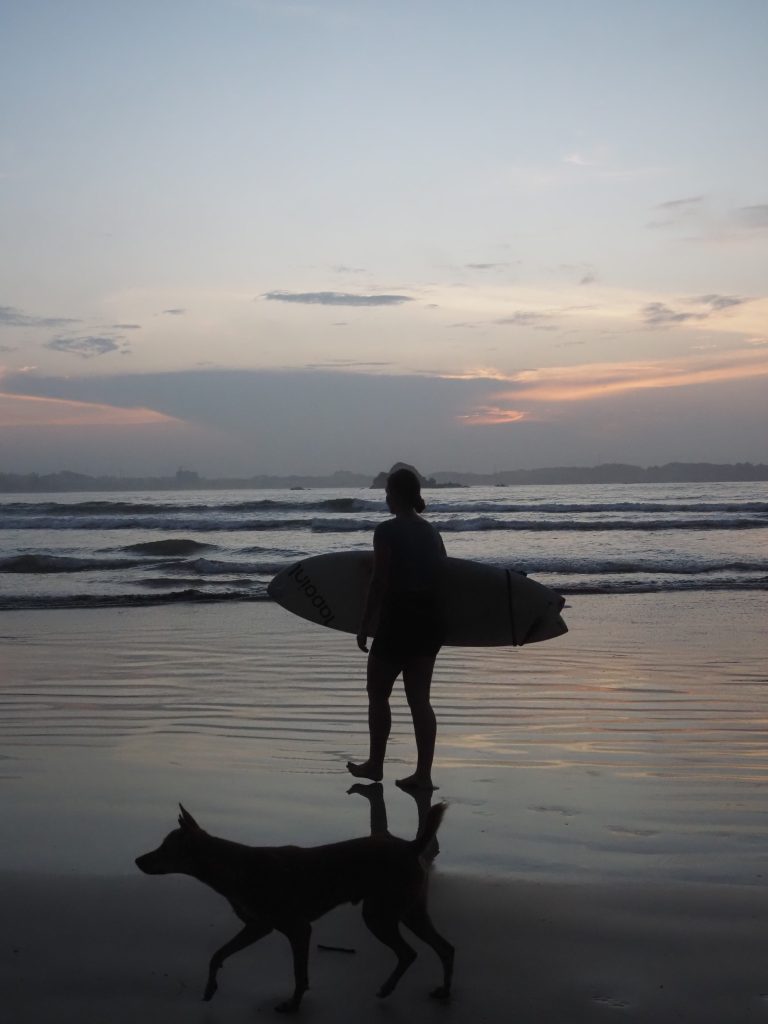 Girl walking with her surfboard and a dog on the beach during sunrise